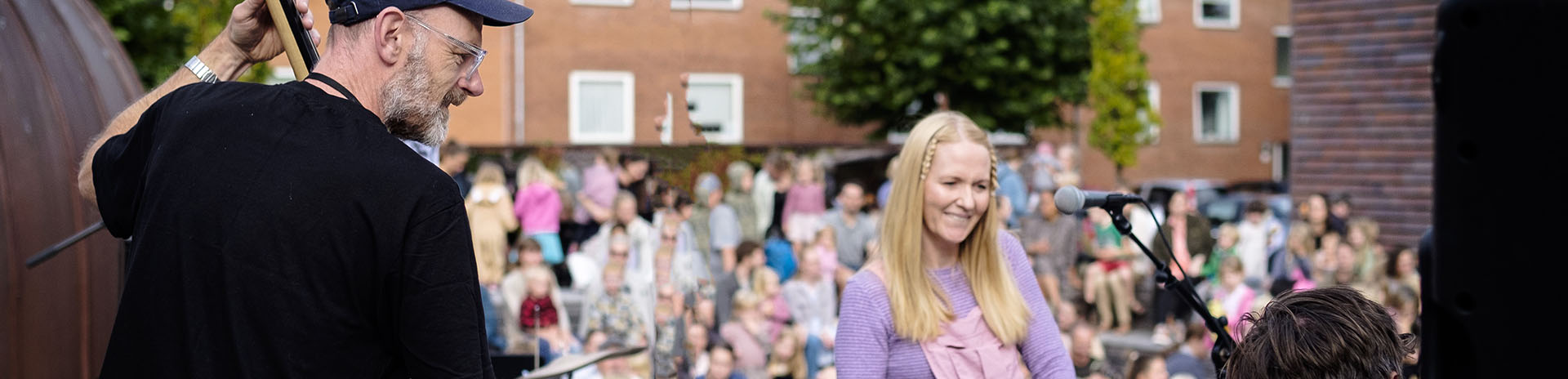 musikalsk optræden udendørs. En mand står til venstre og spiller på en kontrabas, iført en sort t-shirt og en blå kasket. Han er i færd med at stemme eller justere instrumentet. I midten af billedet står en kvinde med langt blondt hår, iført en lyserød buksedragt med en lilla trøje. Hun smiler og ser ud til at være klar til at synge eller tale i mikrofonen foran hende. Til højre i billedet sidder en person ved et keyboard, der ser ud til at være dybt koncentreret. I baggrunden er der en stor forsamling af mennesker, der sidder og står, tilsyneladende som publikum til optrædenen. Bygninger med røde tegltage og træer kan også ses i baggrunden.