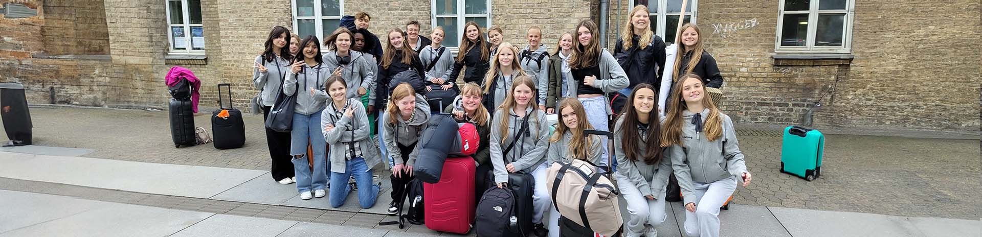 Alle piger er stillet op på Sorø station i et samlet gruppe billede alle smilende forventningsfulde med kufferter og rygsække og grå sponsor hættetrøjer